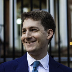 A man in a blue suit and teal tie smiles while standing outdoors in front of a black iron fence and a blurred brick building.