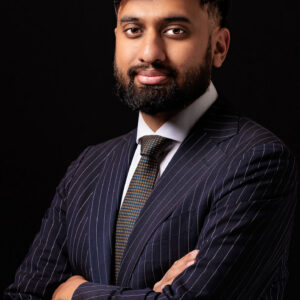 A man with a beard and mustache, wearing a dark pinstripe suit and tie, stands against a black background with arms crossed, looking confidently at the camera.