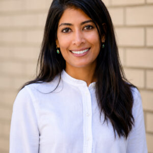 A woman with long dark hair, wearing a white button-up shirt, smiles while standing in front of a light tan brick wall.