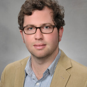 A man with curly hair and glasses wearing a tan blazer and a blue-and-white striped shirt.