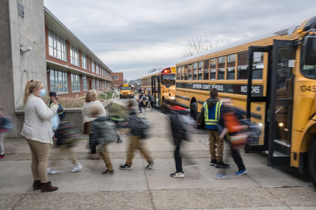 Children wearing backpacks get off yellow school buses and walk toward a school building while adults supervise. The scene is busy, with motion blur showing the students movement. The sky is overcast.
