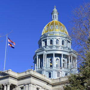 The gold dome of the Colorado State Capitol building against a clear blue sky, with the U.S. and Colorado state flags waving nearby.