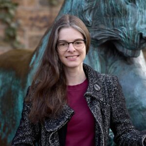 A smiling woman with glasses, wearing a textured jacket over a maroon shirt, stands in front of a bronze statue of a lion.
