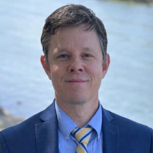 A man in a blue suit, light blue dress shirt, and yellow and blue striped tie stands in front of a body of water.