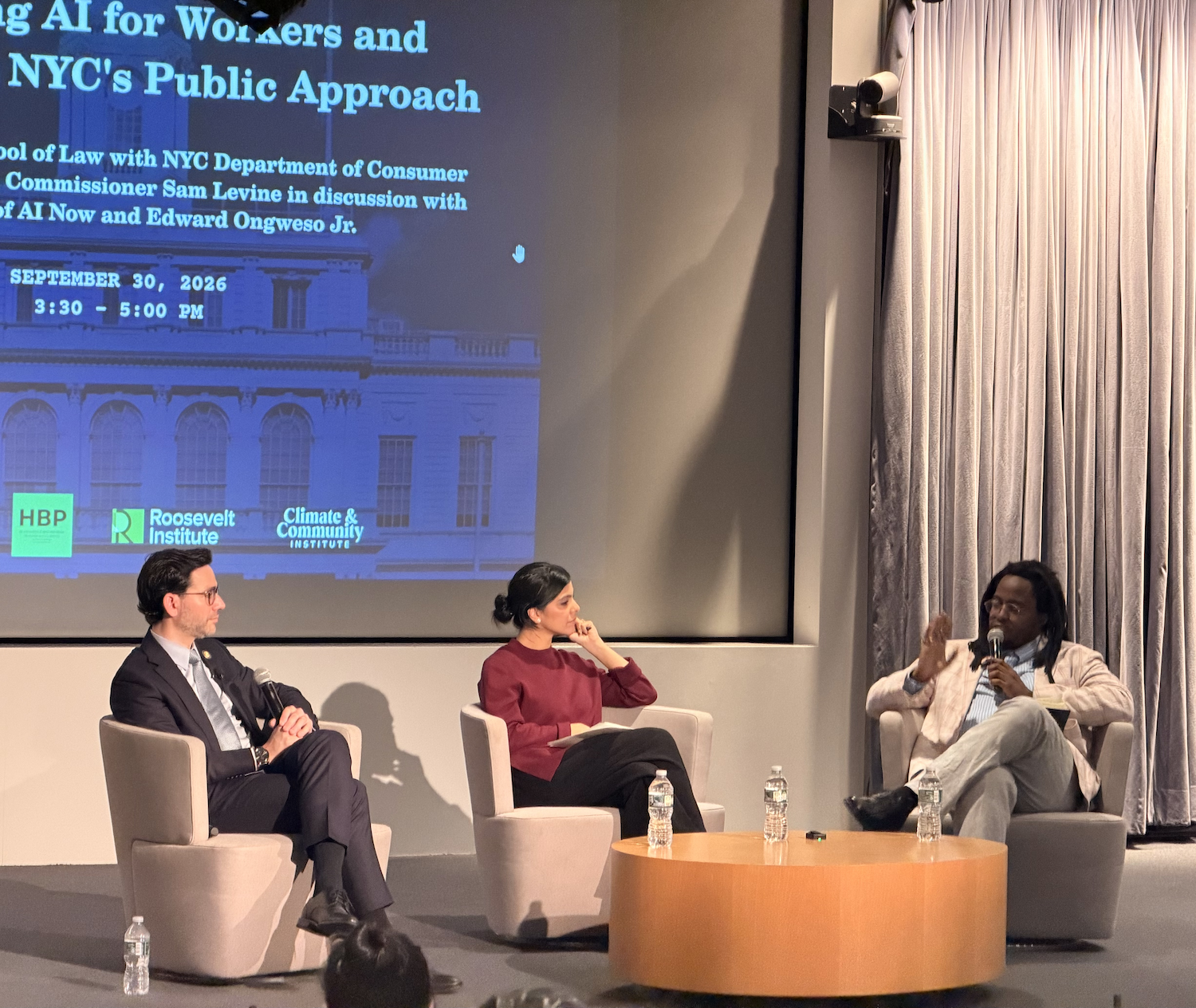 Three panelists sit in armchairs on a stage in front of a screen detailing an event about AI for workers hosted by the Roosevelt Institute and Climate & Community Institute.